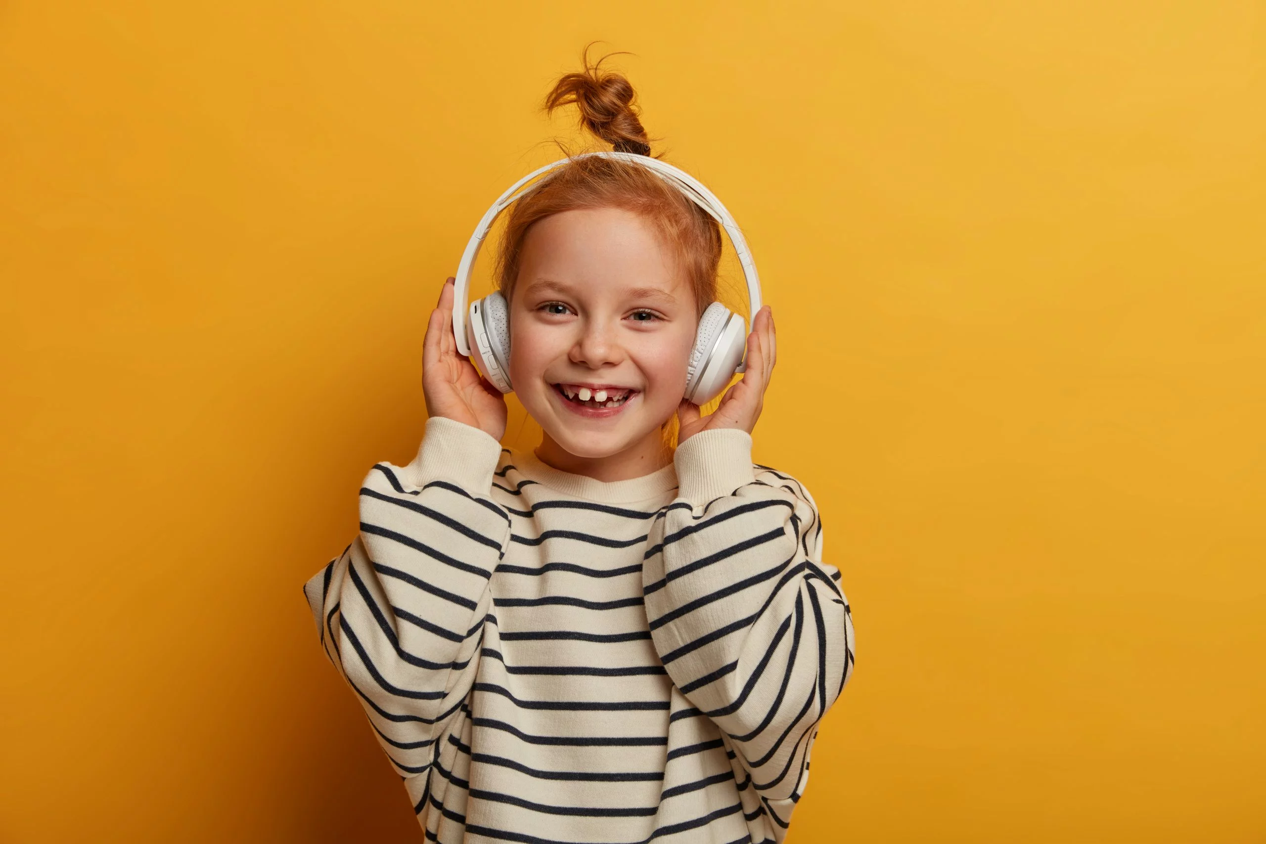 Positive redhead girl enjoys favorite tune, listens music in headphones, has optimistic mood, hair knot, wears striped jumper in casual style, poses against yellow background, smiles toothily